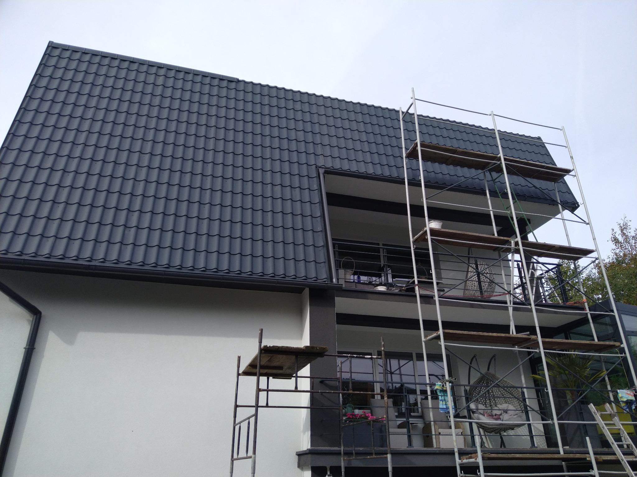 Nowoczesny dom z ciemnoszarą dachówką karpiówką i metalowymi rynnami, częściowo zasłonięty rusztowaniem podczas prac budowlanych na balkonie.