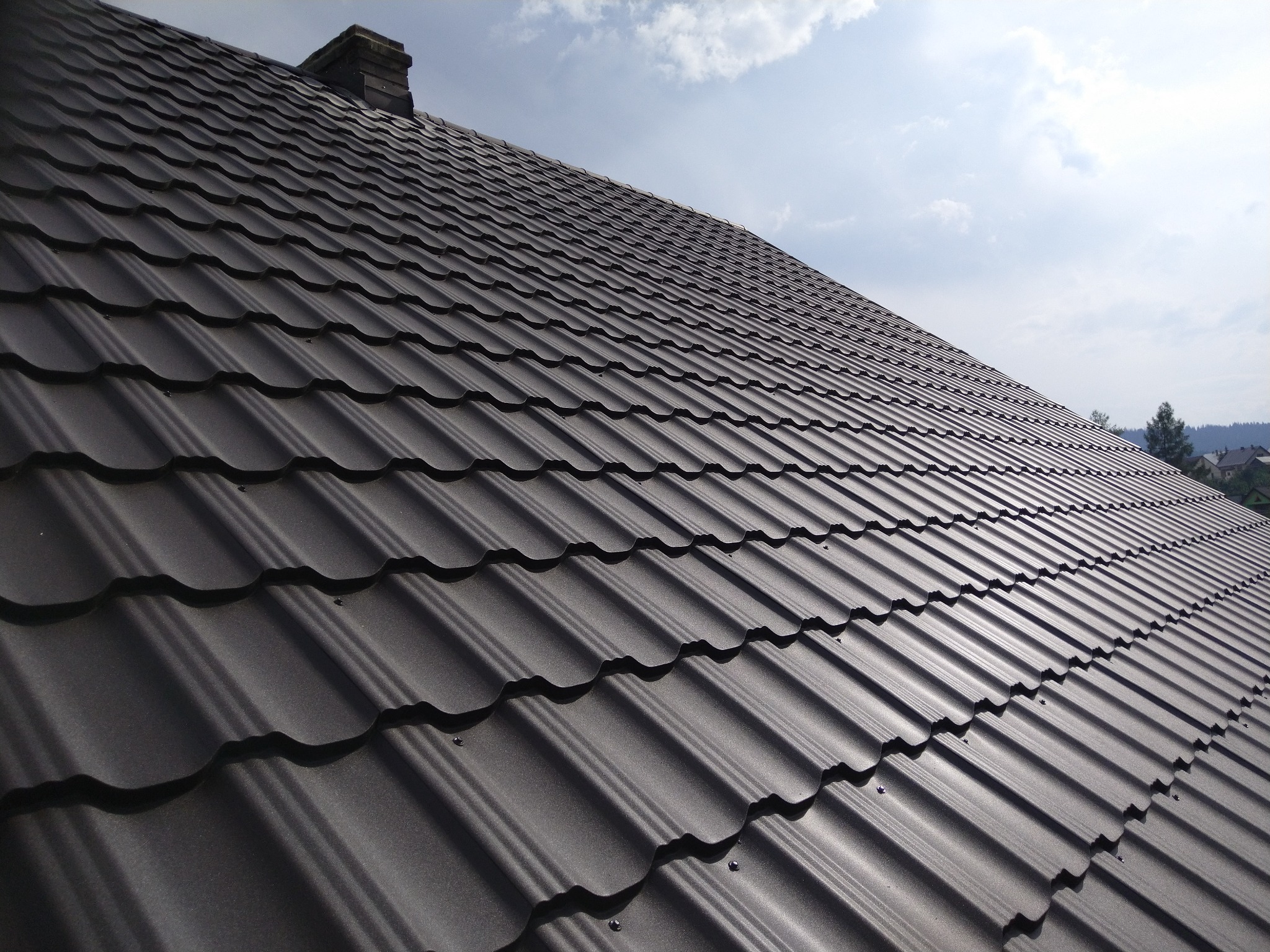 Nowe, ciemnoszare pokrycie dachowe z blachodachówki modułowej, widok z dołu pod kątem, komin w tle, błękitne niebo z chmurami.