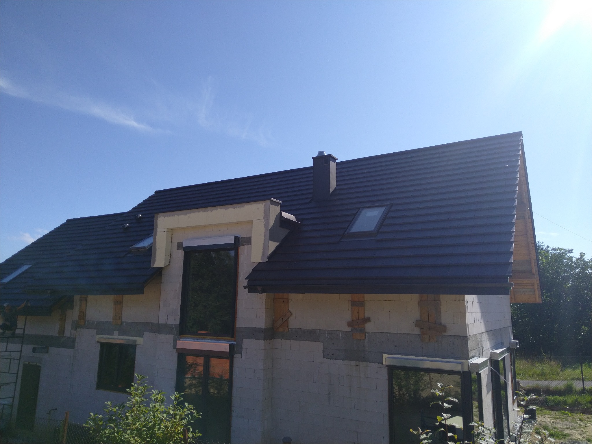 Dom w trakcie budowy z zamontowanym, ciemnym dachem z dachówki, widoczne okna dachowe i komin, ściany z białego bloczka, drewniane elementy szalunkowe, błękitne niebo w tle.