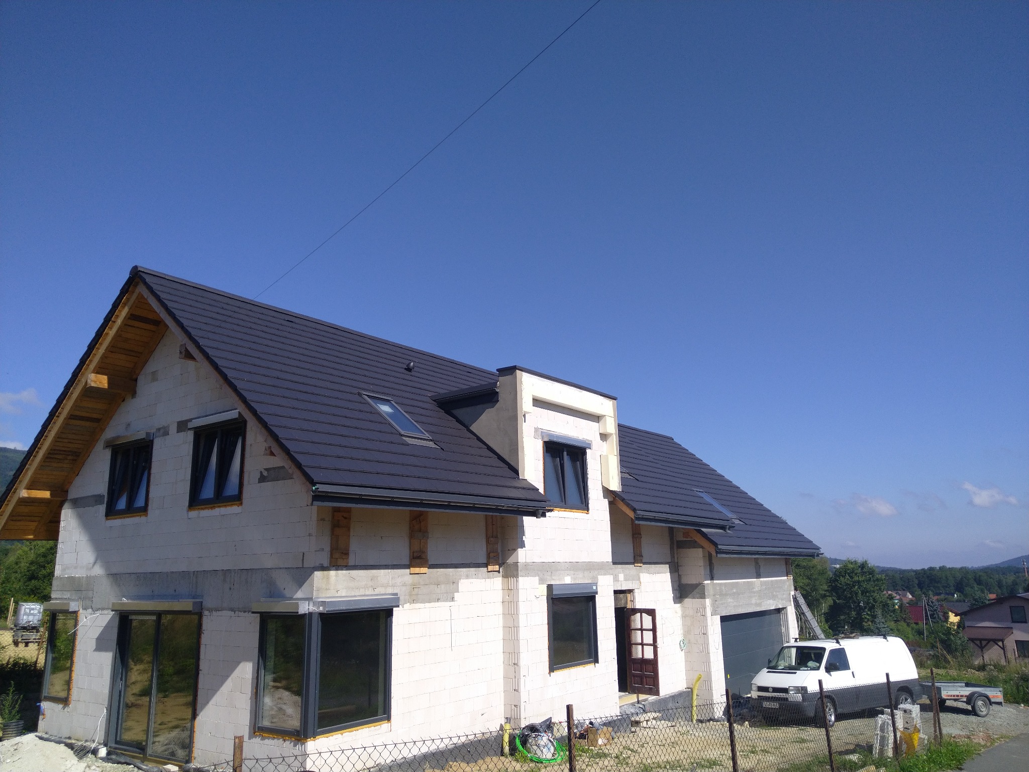 Dom w trakcie budowy z ciemną dachówką i widocznymi oknami, zaparkowany biały bus z przyczepą w tle, ogrodzenie z siatki.