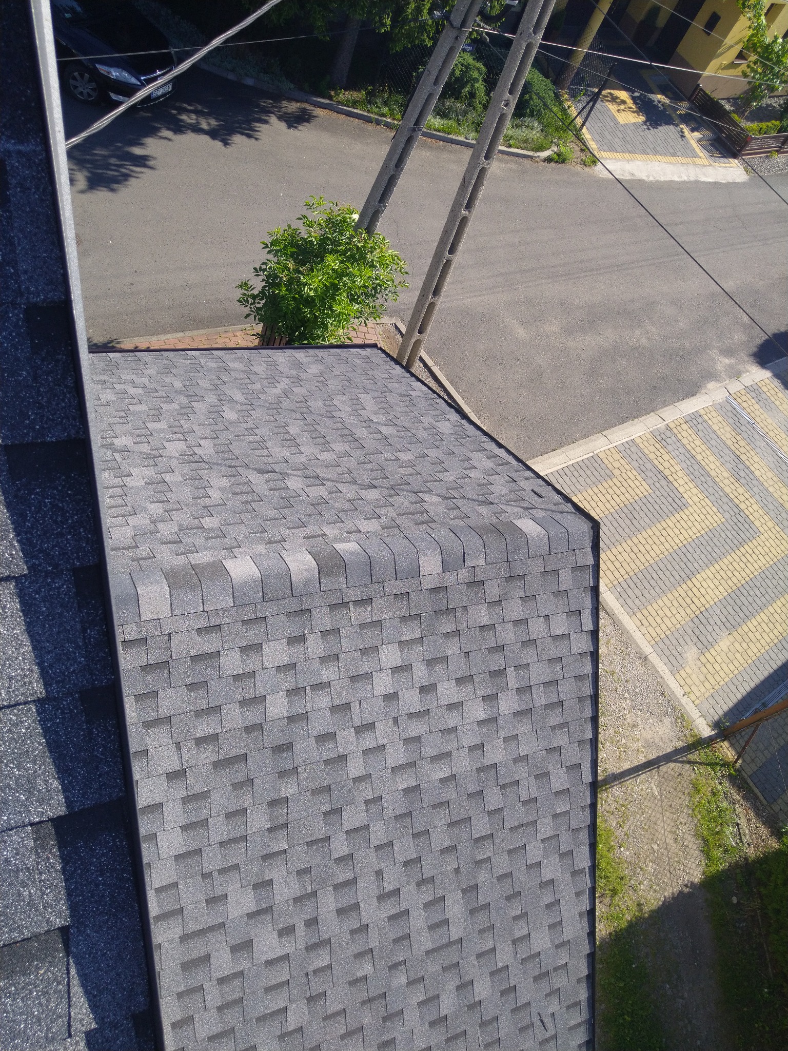 Widok z góry na fragment dachu pokrytego szarym gontem bitumicznym, z widocznym fragmentem rynny i otoczeniem budynku.