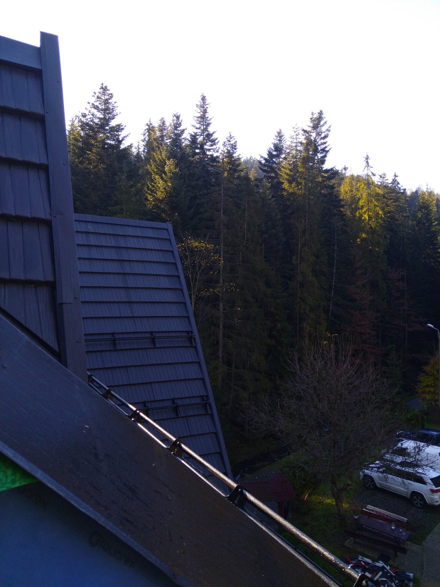 Ciemna, metalowa połać dachu z widocznymi mocowaniami, na tle lasu iglastego i zabudowań z parkującymi samochodami.
