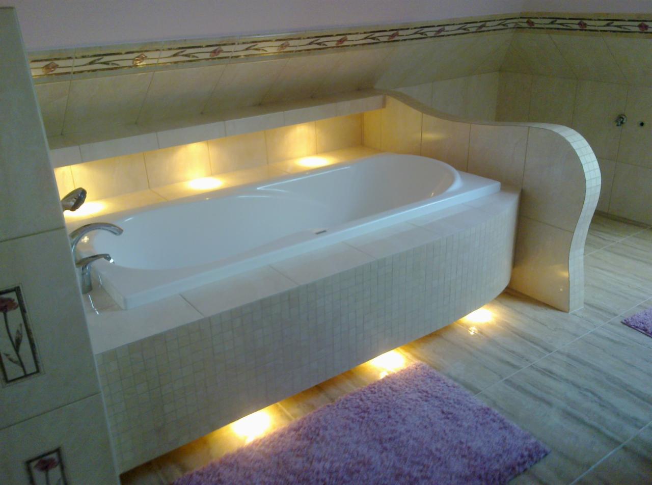 Wanna obudowana mozaiką w łazience na poddaszu z podświetleniem LED, widoczny fragment umywalki z motywem kwiatowym i fioletowy dywanik.