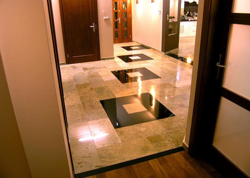 Polerowana podłoga z kamiennych płytek w odcieniach beżu i brązu z czarnymi kwadratowymi intarsjami, widok z perspektywy wejścia, ciepłe oświetlenie.