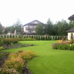 Zadbany ogród z równo przystrzyżonym trawnikiem, kwitnącymi rabatami i żywopłotem z iglaków, widok na dom z jasną elewacją i drewnianymi oknami.