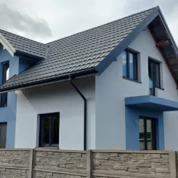 Nowoczesny dom jednorodzinny z elewacją w odcieniach szarości i błękitu, z ciemną dachówką i stolarką okienną, widziany zza betonowego ogrodzenia.