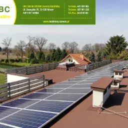 Instalacja paneli słonecznych na płaskim dachu budynku w Mircze, widok z góry na panele, kominy wentylacyjne i fragment zabudowy mieszkalnej w tle.