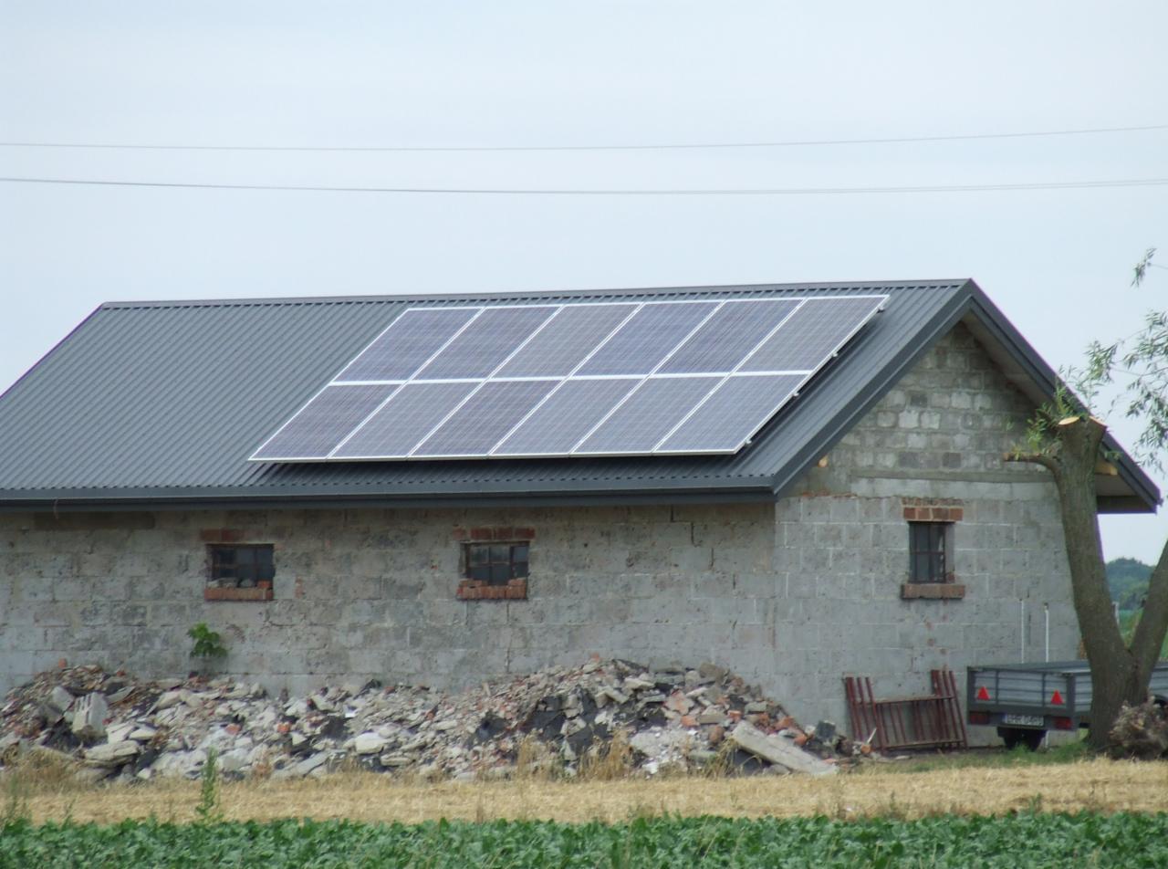 Instalacja paneli słonecznych na szarym dachu budynku gospodarczego z cegły. Widoczne okna, sterta gruzu i przyczepa. Ujęcie w dzień.