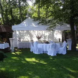 Biały namiot plenerowy rozstawiony na trawniku, z nakrytymi stołami i krzesłami, przygotowany na uroczystość w ogrodzie.