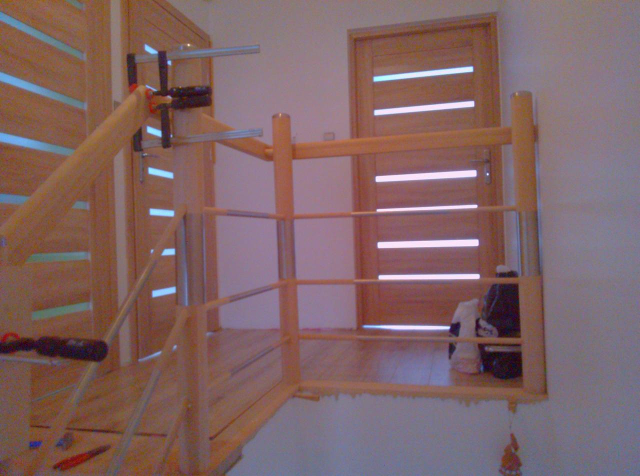Drewniana balustrada schodowa w trakcie montażu, zabezpieczona ściskami stolarskimi, z widocznymi drzwiami z poziomymi przeszkleniami w tle.