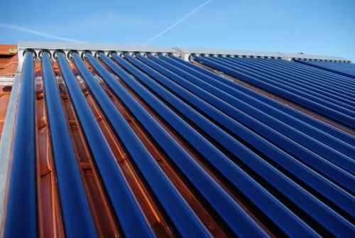 Rząd niebieskich kolektorów słonecznych na dachu z pomarańczowej dachówki, widzianych pod kątem, z jasnym błękitnym niebem w tle.
