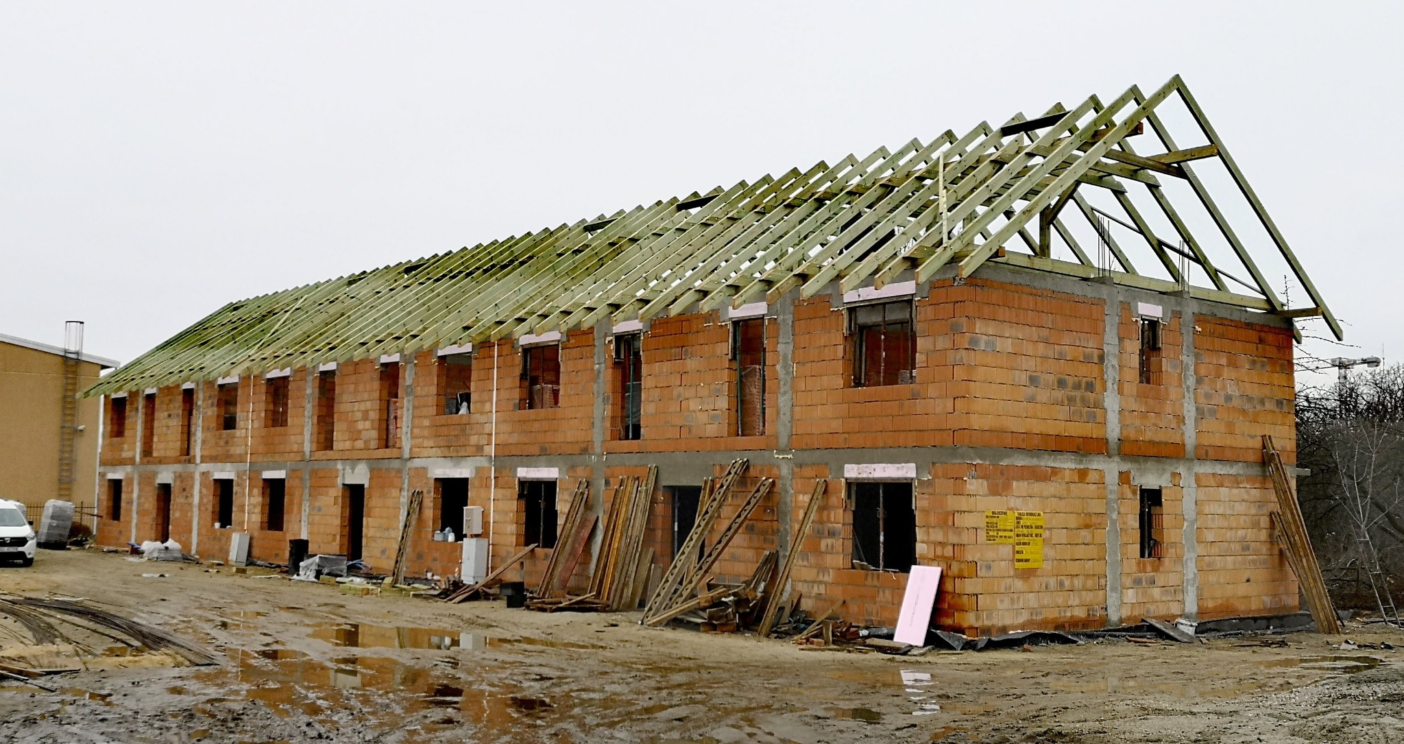 Budowa dwukondygnacyjnego budynku z cegły z widoczną, częściowo ukończoną konstrukcją dachu. Na pierwszym planie błotniste podłoże i materiały budowlane.