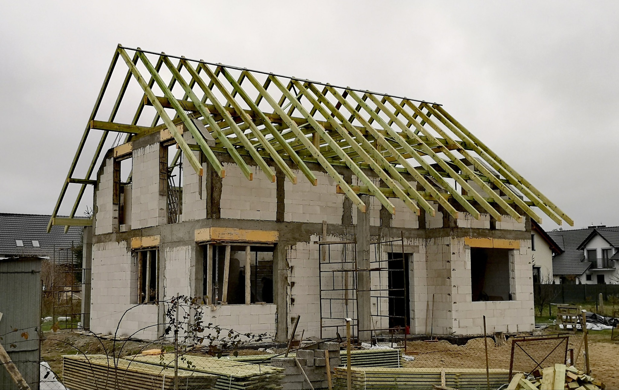 Budowa domu z elementami konstrukcji dachowej w Kotli. Surowy stan, białe ściany, drewniane elementy dachu na tle szarego nieba. Widoczne rusztowania i materiały budowlane.