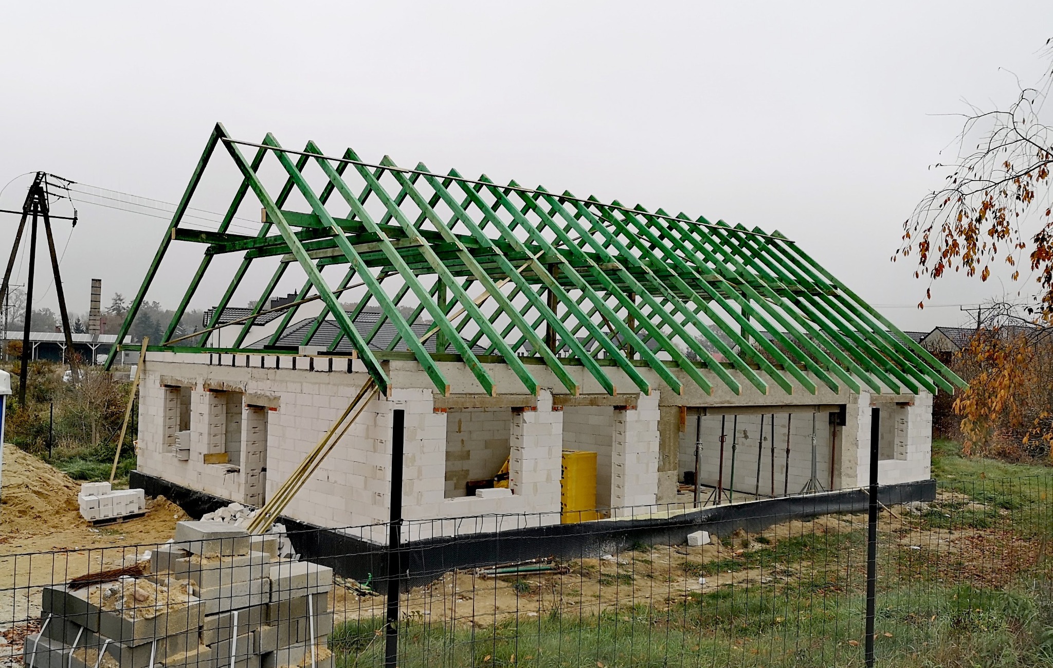 Budowa domu jednorodzinnego w trakcie realizacji. Zielona konstrukcja dachu na ścianach z betonu komórkowego. Widoczne bloczki i teren budowy.