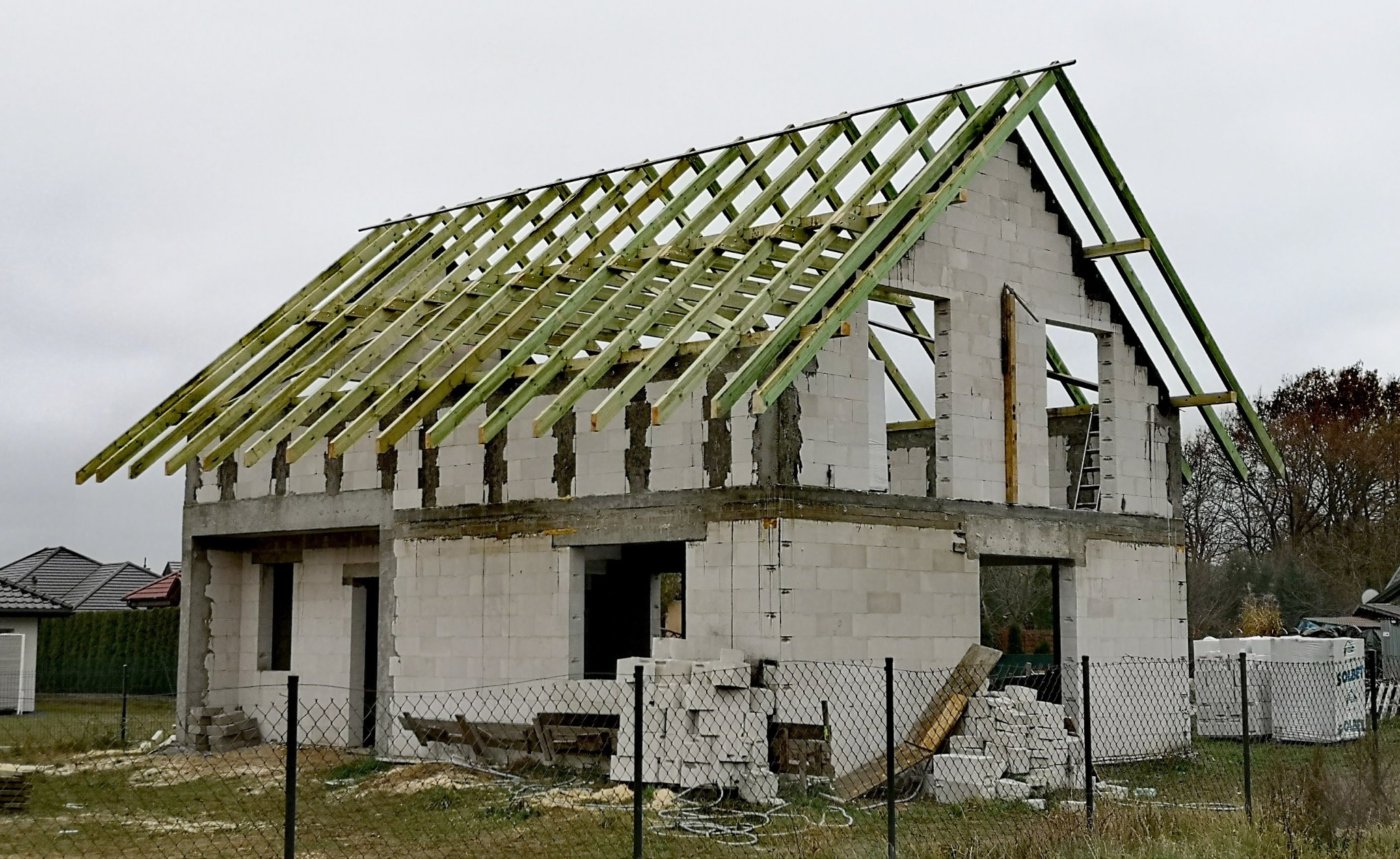 Surowy stan domu jednorodzinnego z widoczną konstrukcją więźby dachowej z impregnowanego drewna, bloki Solbet na paletach. Działka ogrodzona siatką.