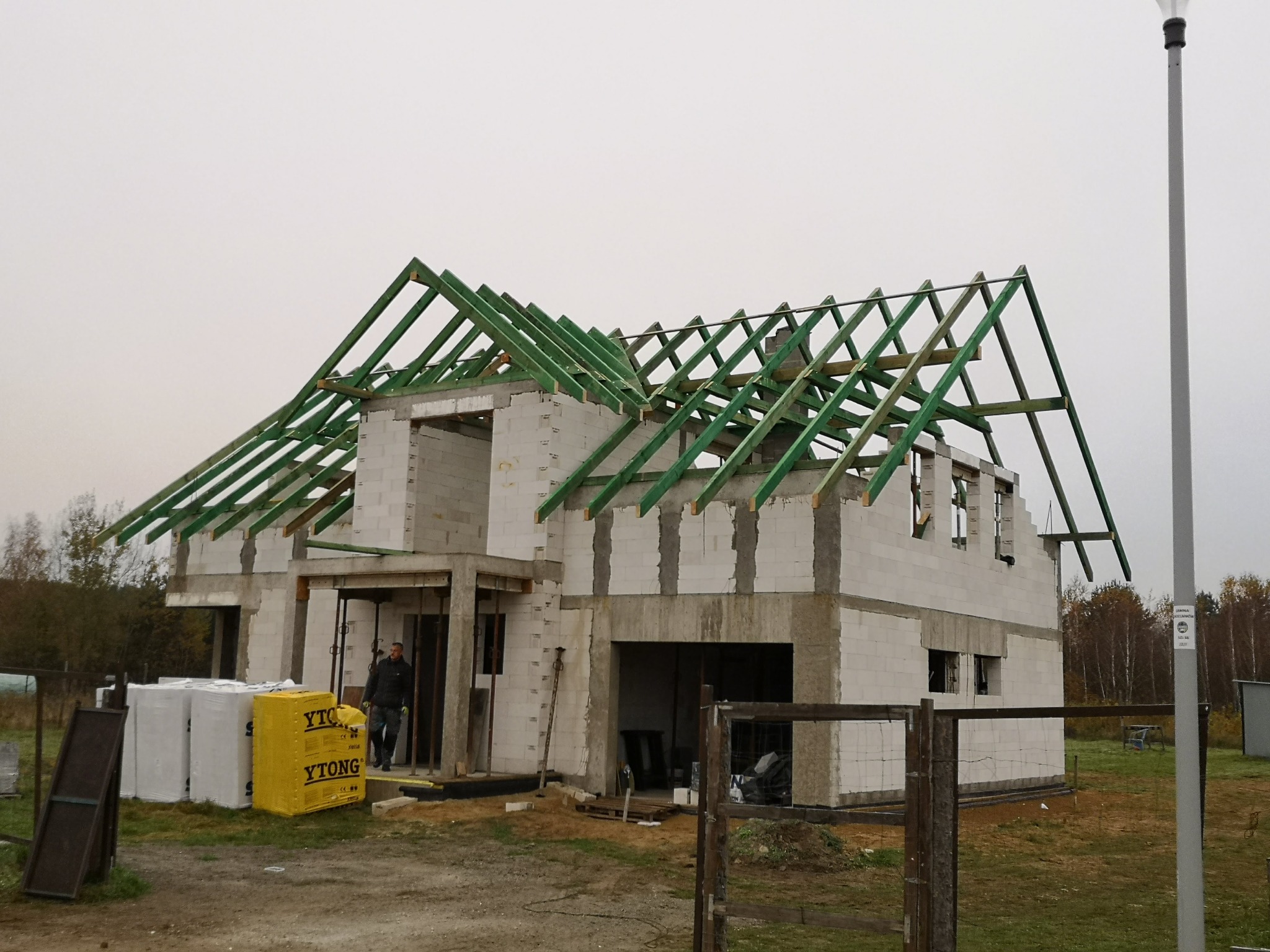 Dom w trakcie budowy z zieloną konstrukcją dachu i blokami YTONG. Widoczny pracownik w drzwiach. Działka z ogrodzeniem i lampą uliczną w tle.
