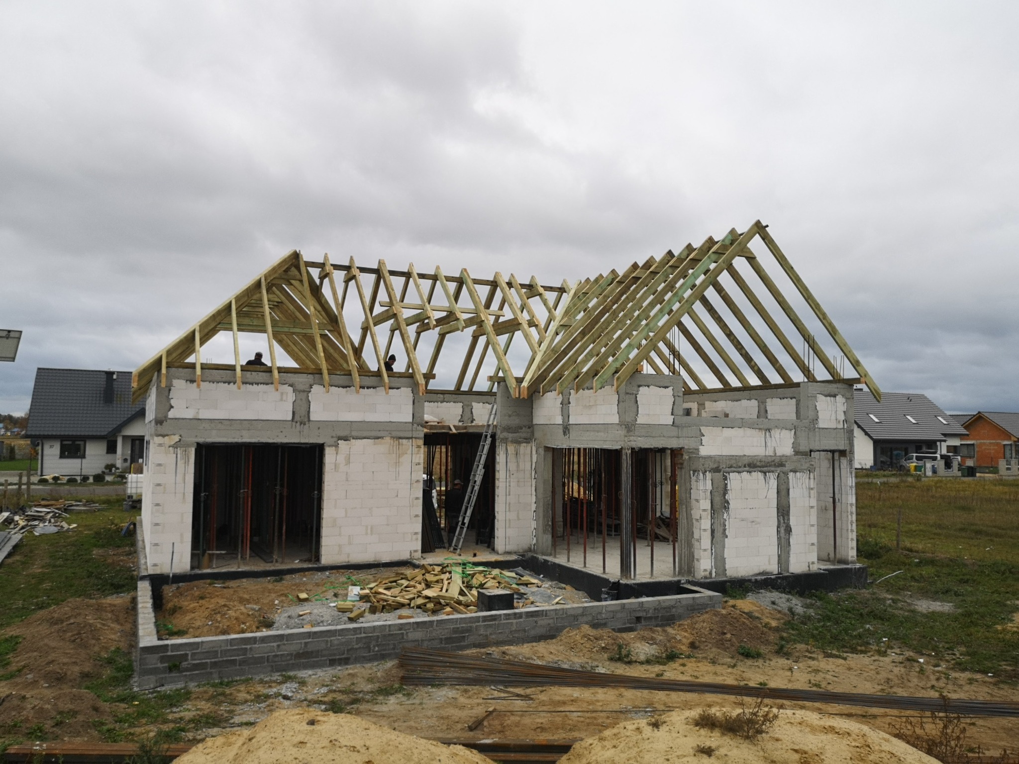 Budowa domu jednorodzinnego: surowy stan z wymurowanymi ścianami i konstrukcją drewnianą dachu w trakcie montażu, widok z zewnątrz.