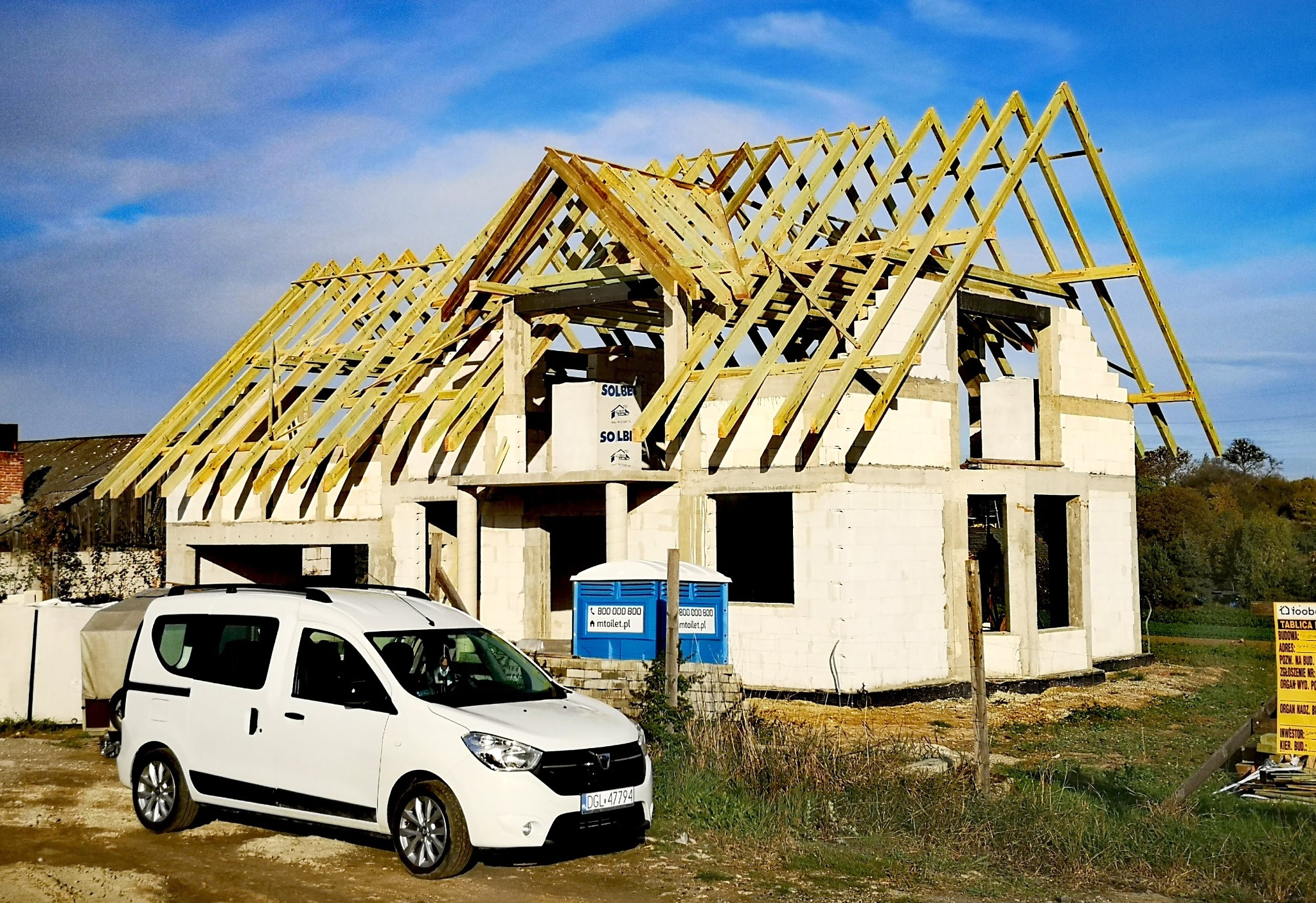 Surowy stan domu z więźbą dachową w trakcie budowy, na tle błękitnego nieba. Na pierwszym planie biały samochód i przenośna toaleta. Widoczna tablica informacyjna budowy.