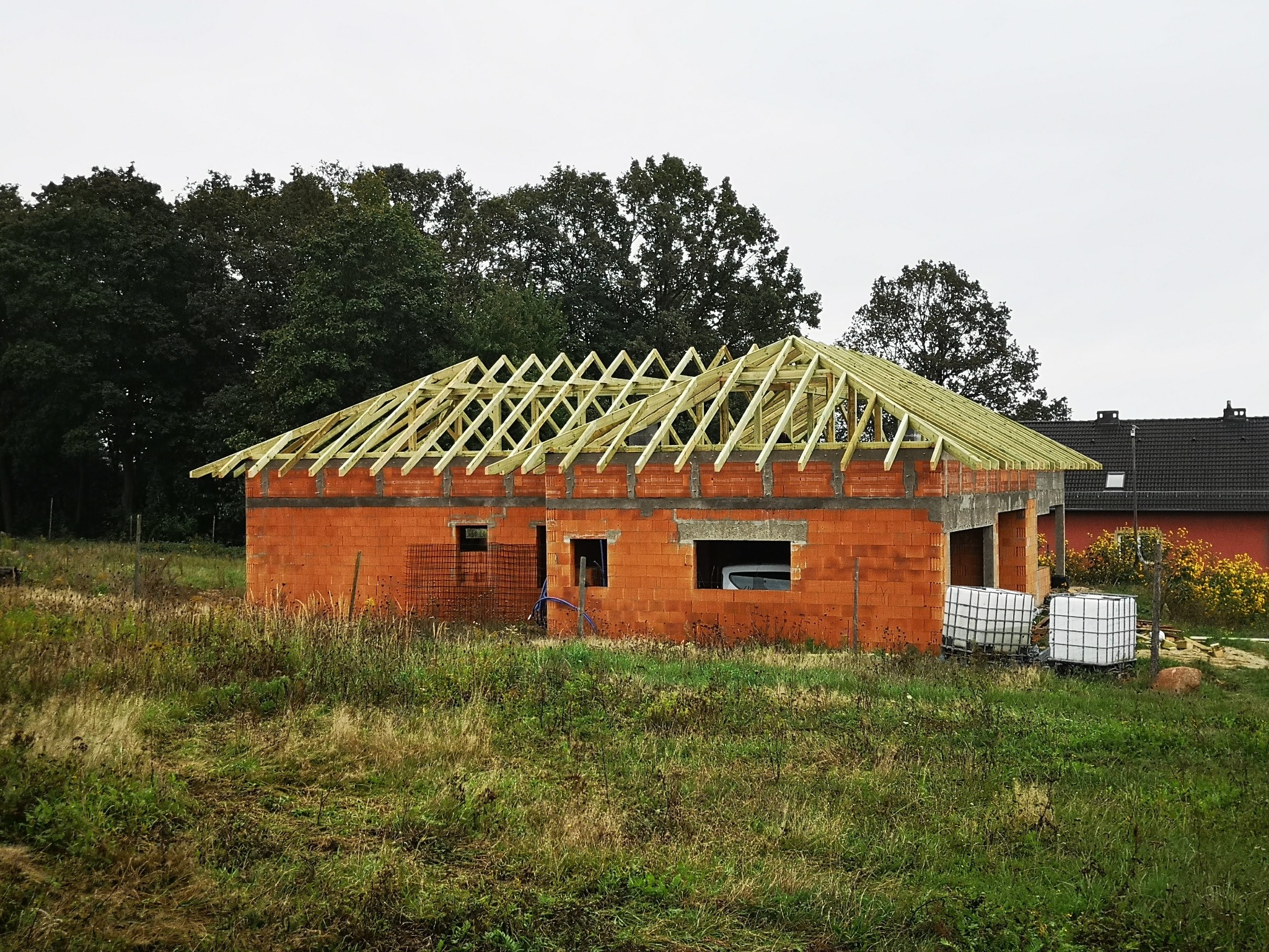 Budowa domu jednorodzinnego w stanie surowym otwartym: ceglany mur i drewniana konstrukcja dachu na tle zieleni i pochmurnego nieba.