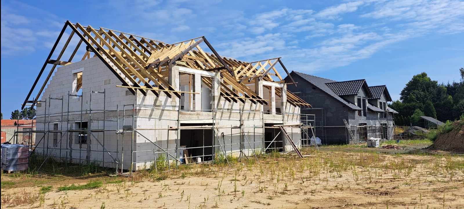 Budowa bliźniaka: surowy stan z widoczną konstrukcją dachu z drewna. Elementy szalunku przy oknach. W tle dom w stanie deweloperskim i błękitne niebo.