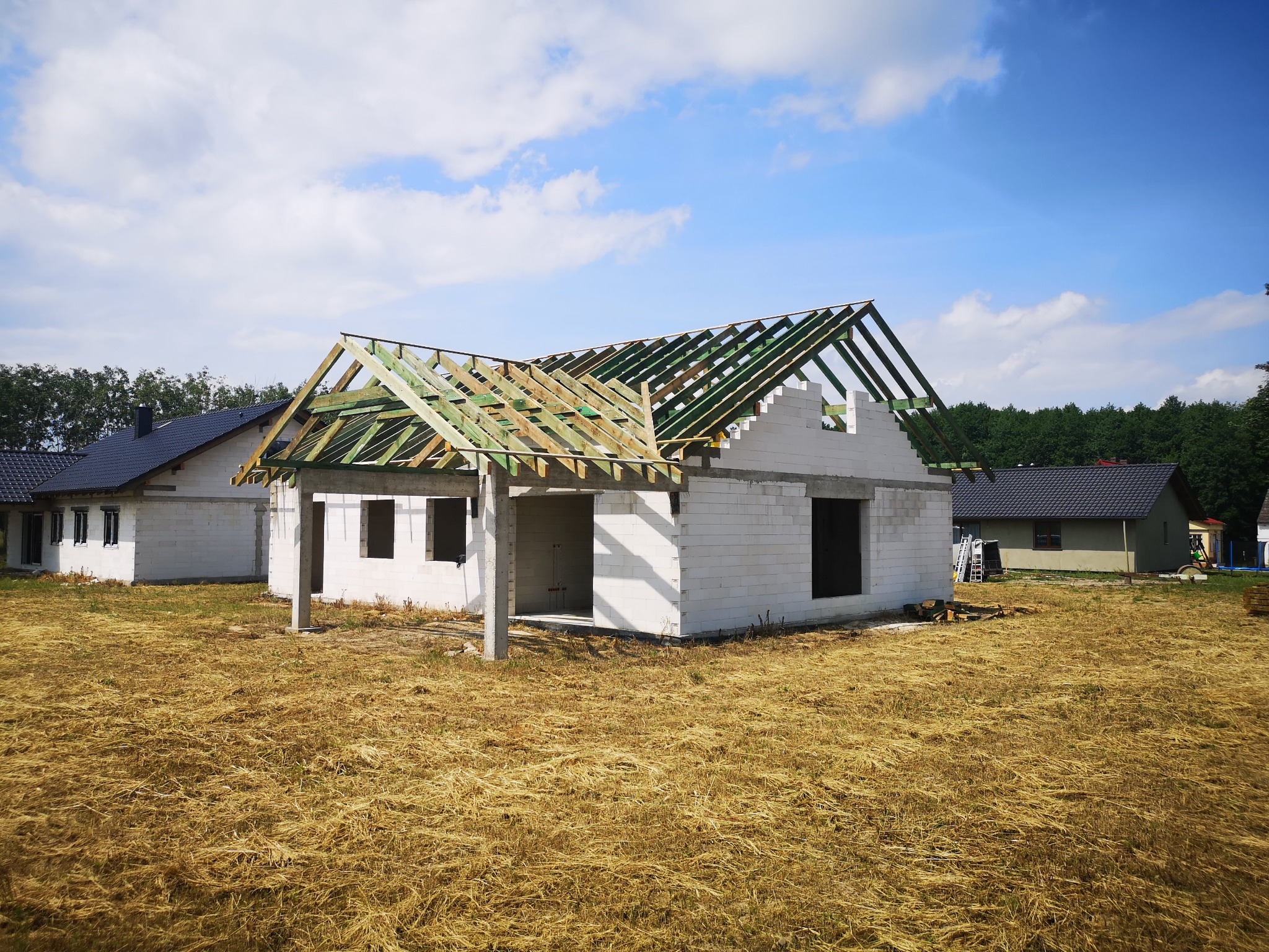 Budowa domu jednorodzinnego w stanie surowym otwartym z widoczną więźbą dachową zaimpregnowaną na zielono, na tle błękitnego nieba.
