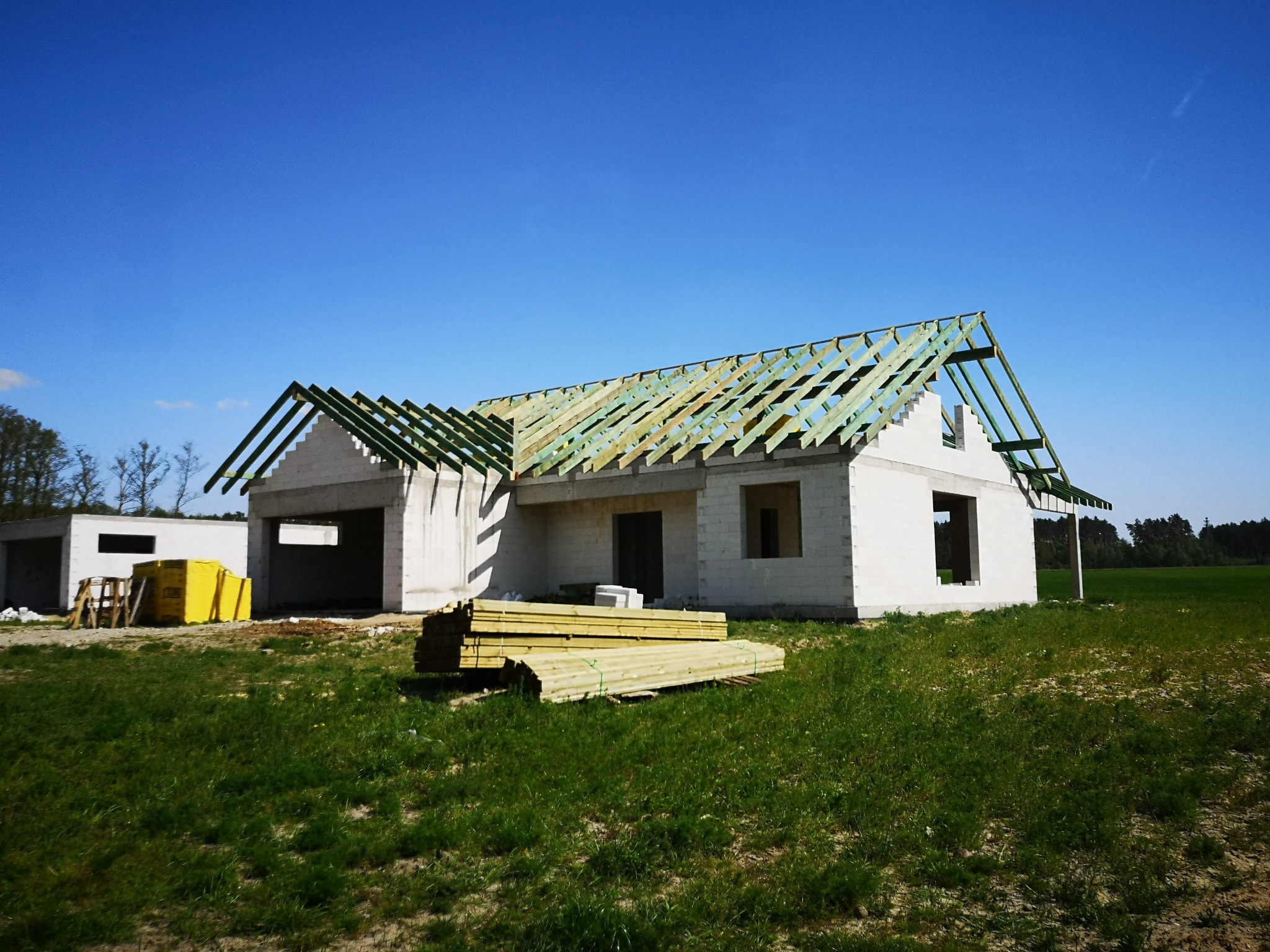 Surowy stan domu w budowie z częściowo zamontowaną, zieloną konstrukcją dachu na tle błękitnego nieba i zielonej trawy. Na pierwszym planie stos drewna.