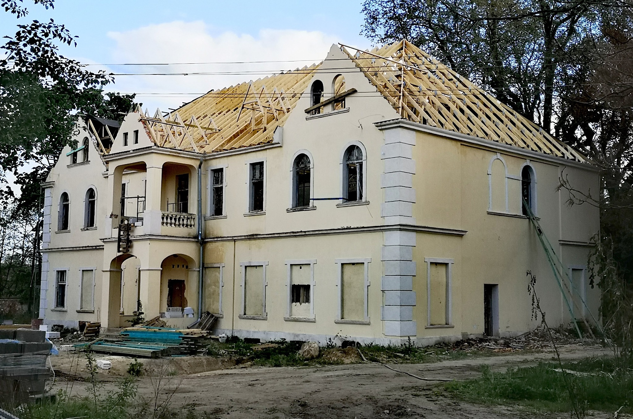 W trakcie budowy: drewniana konstrukcja dachu na tle kremowego, dwupiętrowego budynku z elementami klasycystycznymi. Widoczne rusztowania i materiały budowlane.