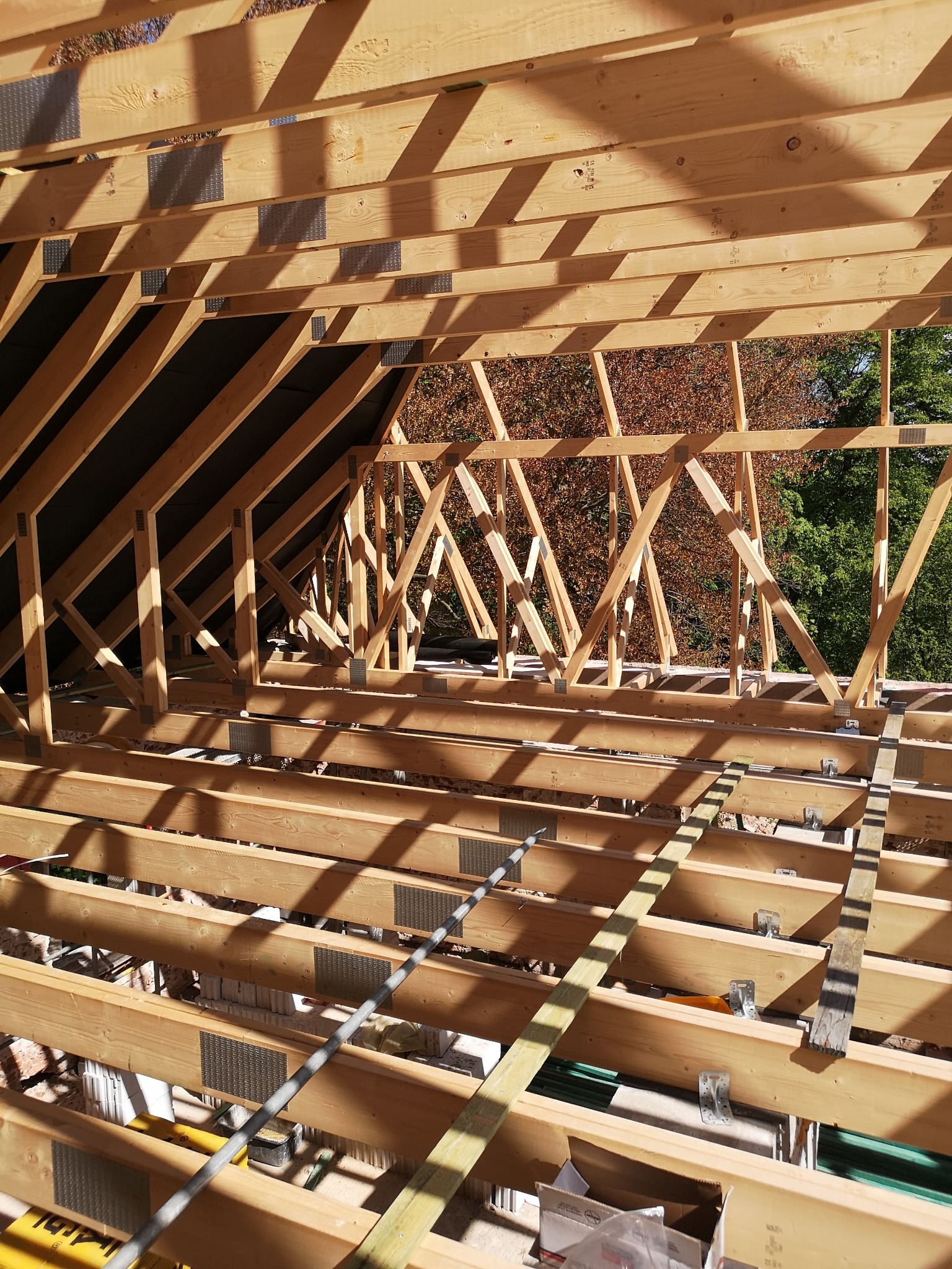 Konstrukcja drewniana dachu z widocznymi elementami łączeń i fragmentami drzew w tle, ukazująca etap budowy z perspektywy wnętrza budynku.