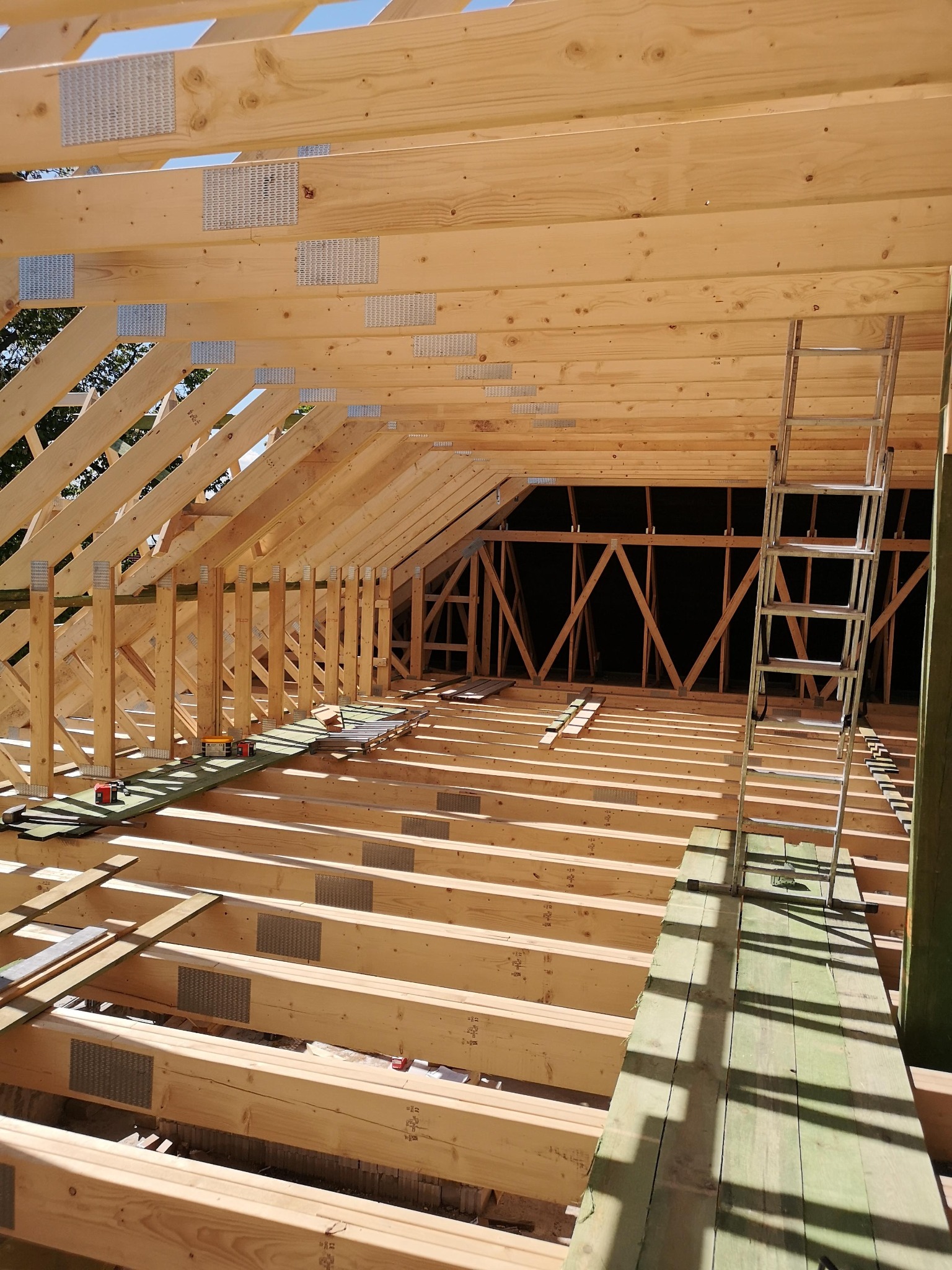 Jasna, drewniana konstrukcja dachu w trakcie budowy, widok z wnętrza. Metalowe łączniki, drabina oparta o ścianę. Widoczne narzędzia i materiały budowlane.