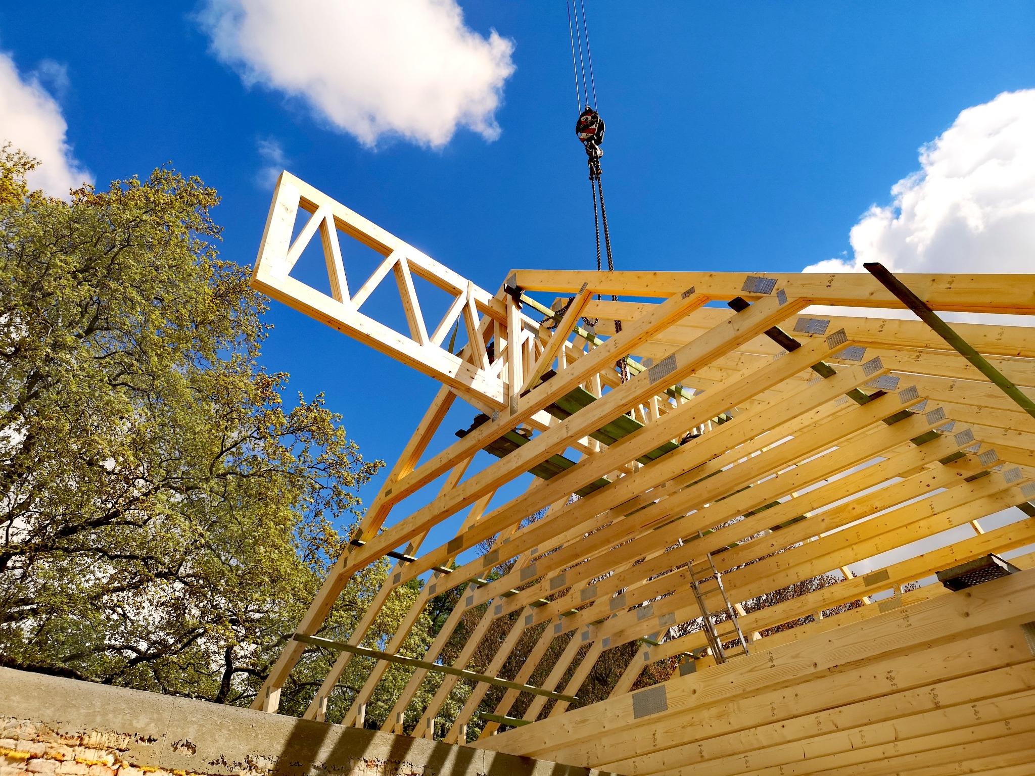 Montaż drewnianej konstrukcji dachowej z użyciem dźwigu na tle błękitnego nieba i drzew. Widoczny fragment murowanej ściany i elementy mocujące.