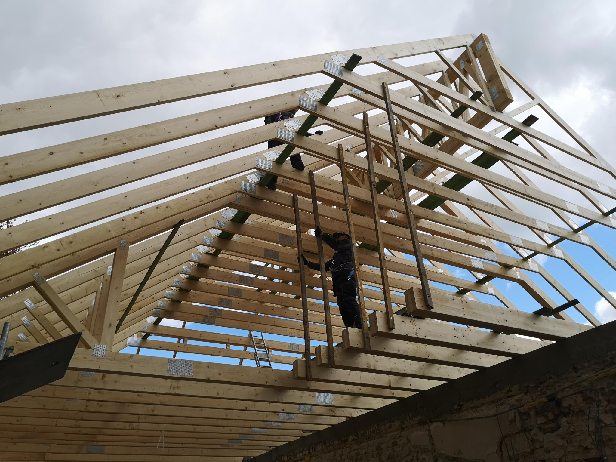 Wznoszenie drewnianej więźby dachowej z widocznymi pracownikami na tle pochmurnego nieba z prześwitami błękitu. Widok z dołu na konstrukcję dachu.