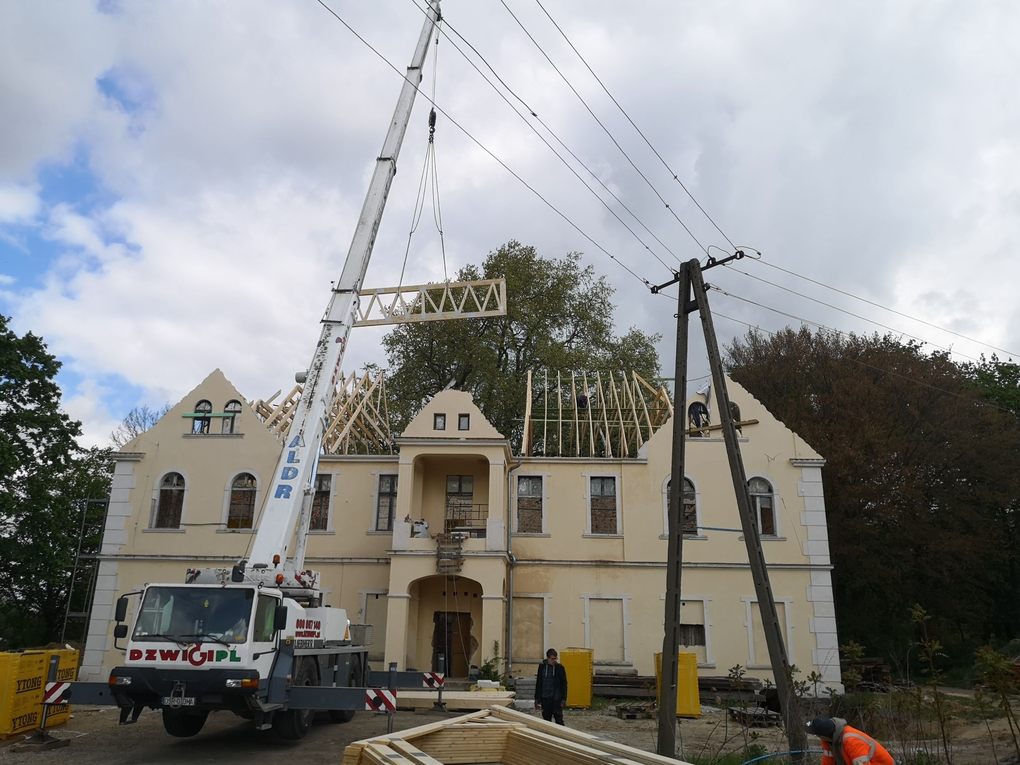 Montaż drewnianej konstrukcji dachowej z użyciem dźwigu na tle budynku z żółtą elewacją. Widoczne elementy więźby i pracownicy w trakcie robót.