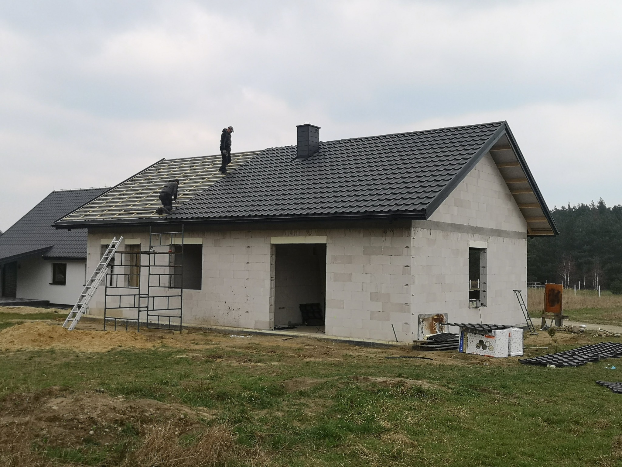 Budowa domu jednorodzinnego: dekarze montują czarną blachodachówkę na drewnianej konstrukcji dachu. Widoczne rusztowania, materiały budowlane i niezadaszona bryła budynku.