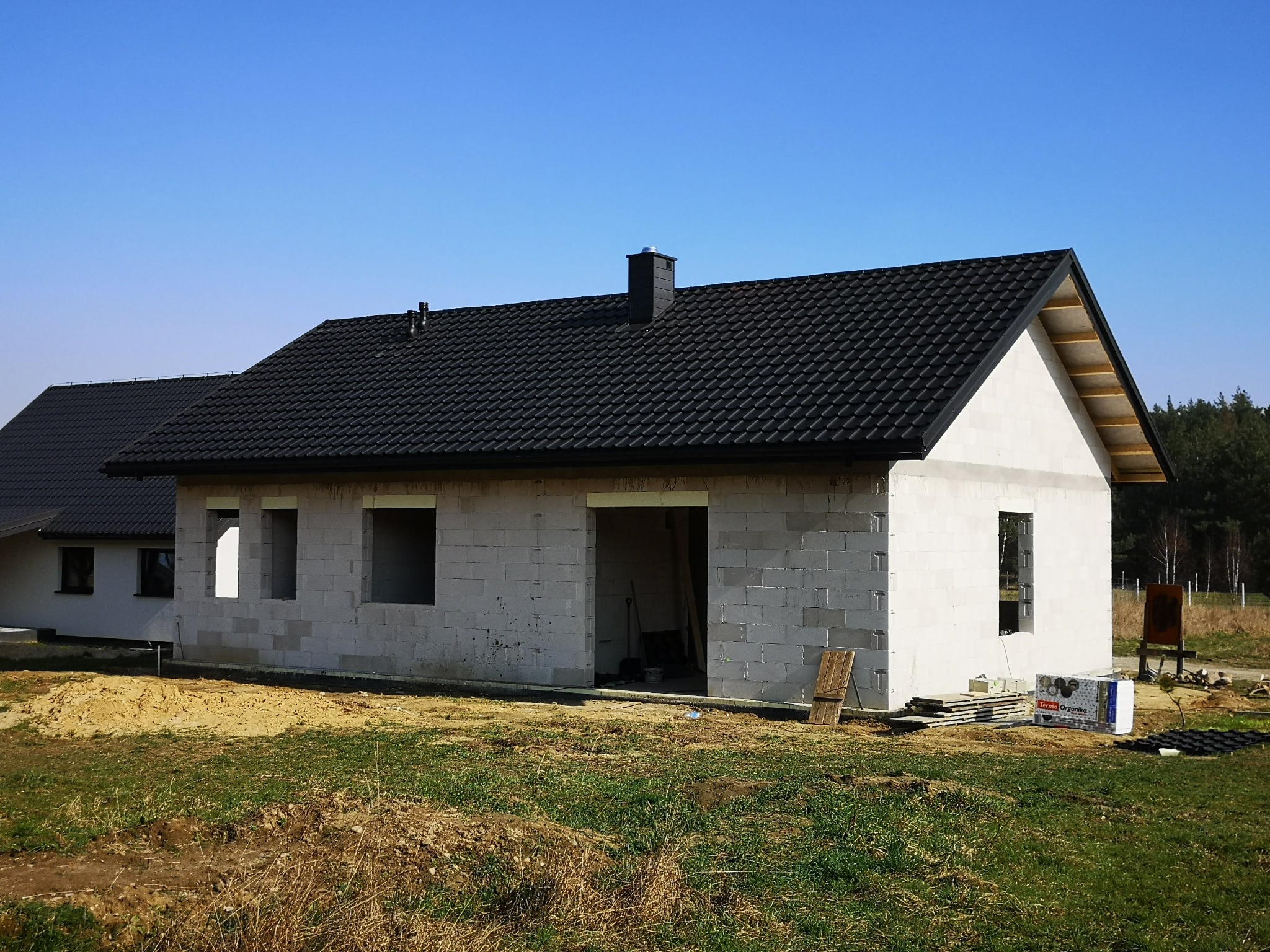Dom w trakcie budowy z czarnym dachem, widoczne bloczki i elementy więźby dachowej. Na trawie leżą materiały budowlane.