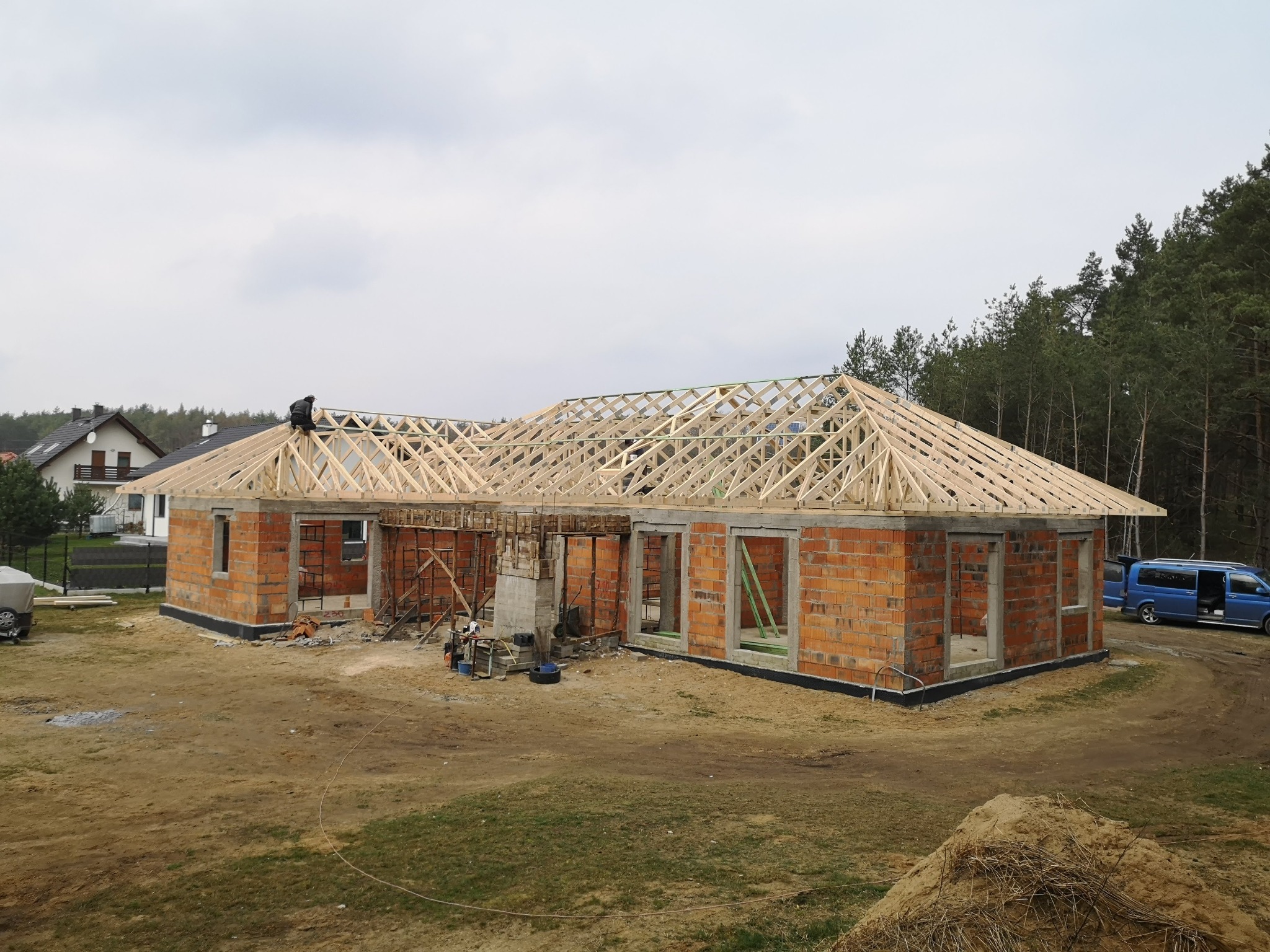 Budowa domu jednorodzinnego w stanie surowym otwartym. Widoczna ceglana konstrukcja ścian i drewniana konstrukcja dachu. Na dachu pracuje dekarz.