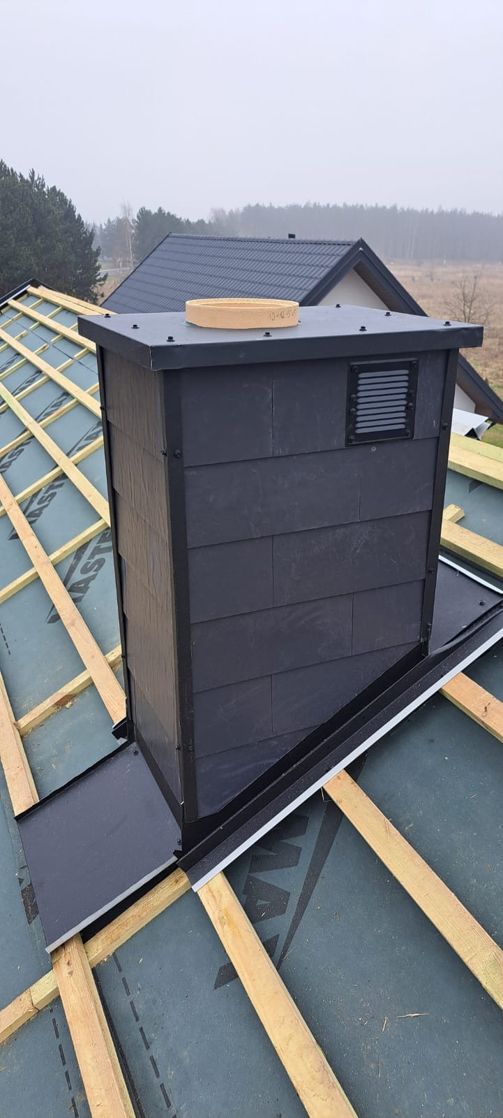 Nowoczesny komin z szarej okładziny na tle drewnianej konstrukcji dachu i zielonej membrany, widok z góry, w tle zabudowa jednorodzinna i drzewa.