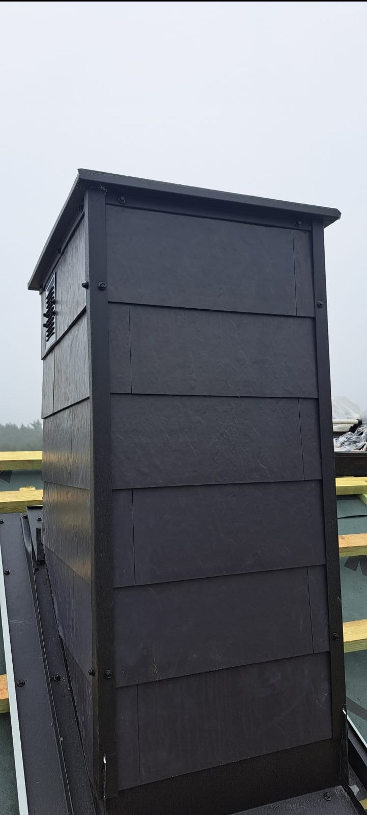 Obróbka komina z grafitowej dachówki łupkowej, zamontowana na dachu z widoczną więźbą. Detal wykończenia komina z bliska w pochmurny dzień.