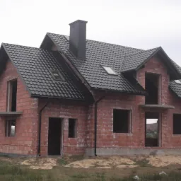 Surowy, dwukondygnacyjny dom z cegły z czarnym dachem z dachówki, widoczne okna dachowe i komin. Budynek w trakcie budowy, bez okien i drzwi.