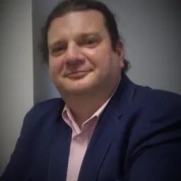 Portret uśmiechniętego mężczyzny w niebieskiej marynarce i różowej koszuli, oparty o szarą ścianę.