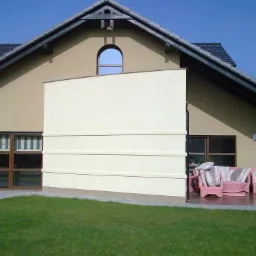 Elewacja domu z beżową ścianą i oknami z brązowymi ramami. Na tarasie różowe meble ogrodowe. Widoczna duża, jasna ściana, prawdopodobnie roleta zewnętrzna.