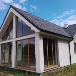 Nowoczesny dom z dużymi oknami PCV w Poznań. Widoczne okna o drewnianych ramach i grafitowy dach. Budynek w stylu minimalistycznym, z wyraźnymi liniami i prostą bryłą.