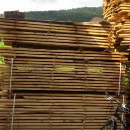 Stosy jasnego drewna tarcicowego zabezpieczone białymi taśmami, z rowerem opartym o jeden ze stosów, na tle zieleni i zalesionego wzgórza pod pochmurnym niebem.