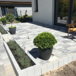 Nowoczesny taras z szarej kostki brukowej ułożonej we wzór, z donicami z roślinami i niskim murkiem rabatowym, na tle białego domu z dużym oknem.