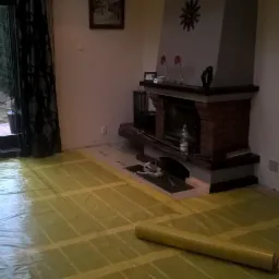 Rozłożona folia grzewcza pod panele w salonie z kominkiem, widoczny fragment instalacji przy kominku oraz rolka folii na podłodze.