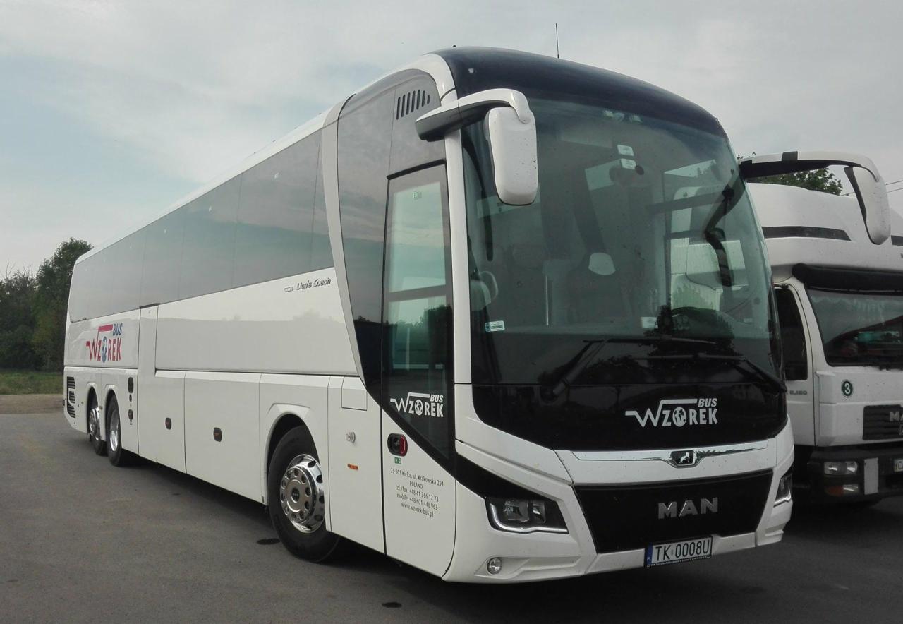 Nowoczesny, biało-czarny autokar turystyczny marki MAN z logo 'WZOREK BUS' na tle pochmurnego nieba i zaparkowanej obok ciężarówki.