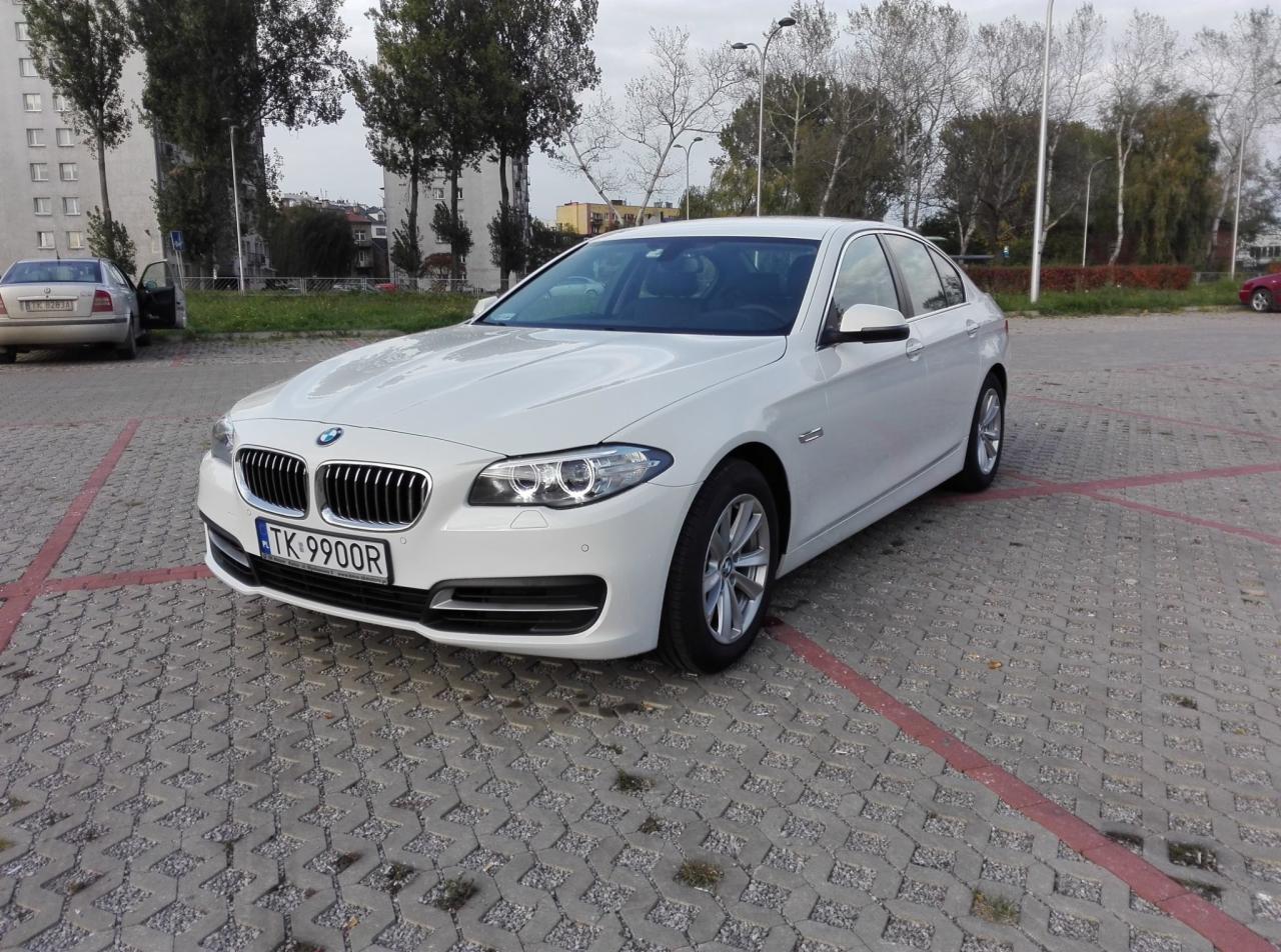 Białe BMW serii 5 na parkingu z kostki brukowej, widoczne tablice rejestracyjne TK 9900R.
