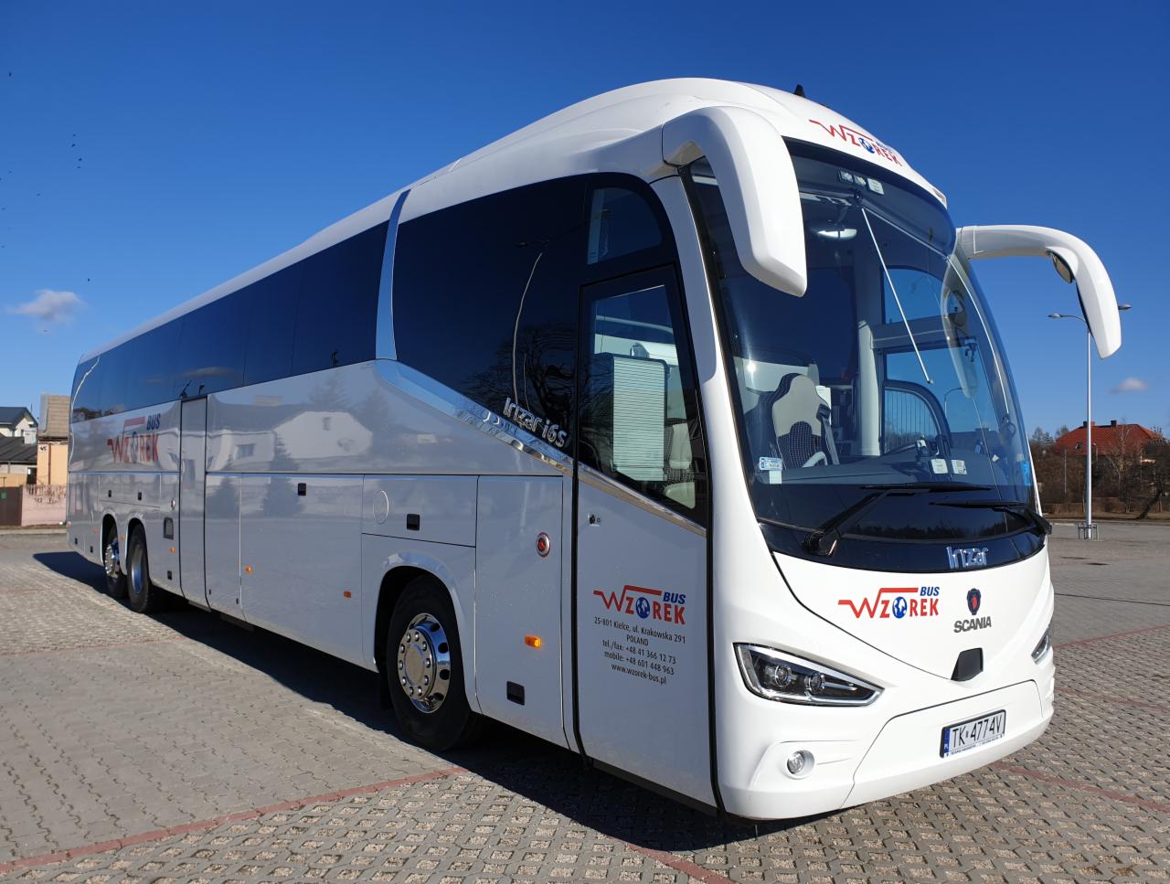 Biały autokar turystyczny Irizar i6s firmy Wzorek Bus zaparkowany na parkingu z kostki brukowej w słoneczny dzień. Widoczne logo firmy i numer rejestracyjny pojazdu.