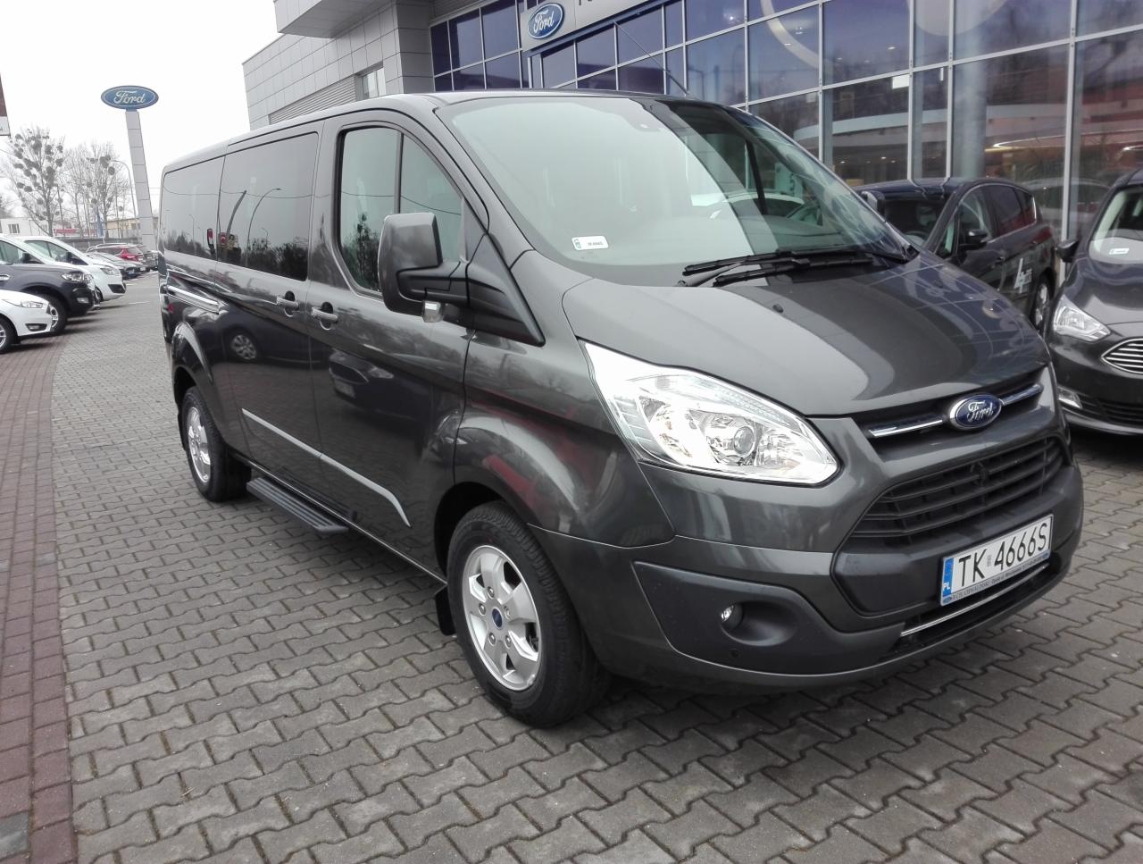 Szary Ford Transit Custom z bocznym stopniem, zaparkowany przed salonem samochodowym Ford.