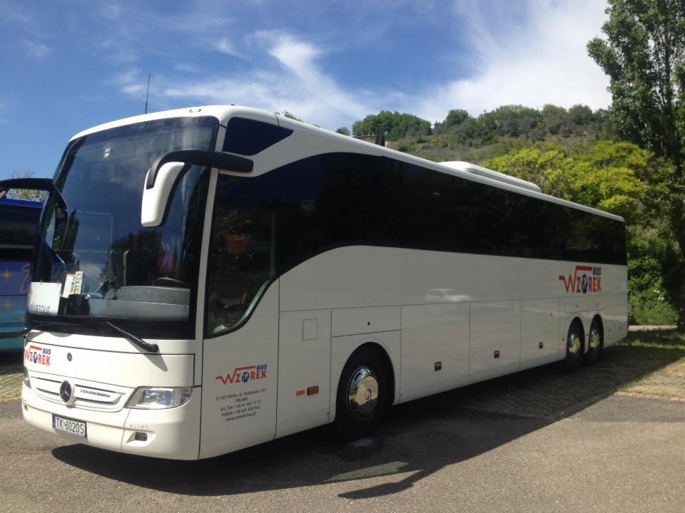 Biały autokar turystyczny z ciemnymi szybami i logo 'Wzorek Bus' na tle zieleni i błękitnego nieba z chmurami.