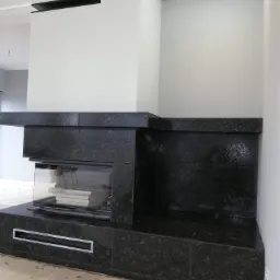 Nowoczesny kominek obłożony ciemnym, błyszczącym granitem, z wkładem kominkowym i półką, w surowym wnętrzu podczas prac wykończeniowych.